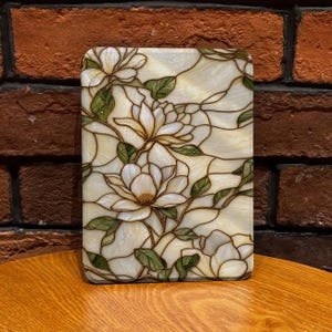 May include: A rectangular stained glass panel featuring a floral design of white magnolia blossoms with yellow centers and green leaves. The glass pieces are joined by dark metal lines, creating a detailed botanical pattern across the surface.