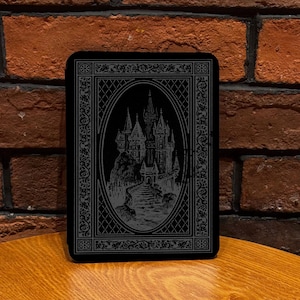 May include: A rectangular black card featuring a detailed silver-toned illustration of a castle with tall spires and a stone path leading to the entrance, enclosed within an oval frame and an intricate decorative border with floral patterns.