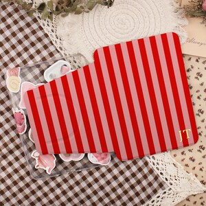 May include: A rectangular phone case featuring a vertical striped pattern in alternating shades of bright red and soft pink. The bottom right corner of the case displays the gold-coloured capital letters JT. The case is transparent with various cake-shaped stickers visible underneath.