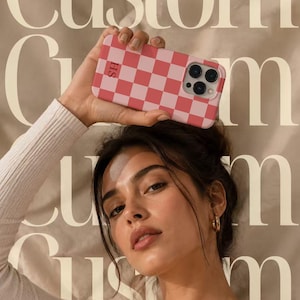 May include: A rectangular smartphone case featuring a pink and white checkered pattern. The case includes a camera cutout on the upper right side and the letters SH printed in a small, dark font on the upper left side of the checkered surface.