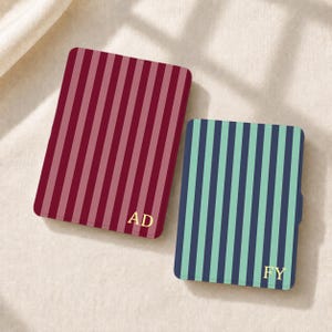 Puede incluir: Dos estuches rectangulares con rayas verticales. El estuche más grande tiene rayas granates y rosa oscuro con las letras doradas AD. El más pequeño tiene rayas azul marino y verde claro con las letras doradas FY. Ambos tienen esquinas redondeadas.