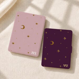 Pode incluir: Duas capas retangulares para e-reader, uma em rosa claro e outra em roxo escuro, ambas com um padrão de folha de ouro de luas crescentes e estrelas. A capa rosa é personalizada com as letras brancas HS e a roxa com as letras VU.