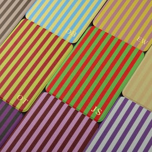 May include: A selection of rectangular coasters with vertical stripes in a variety of colours, such as red, green, yellow, blue, and purple. Some coasters have gold initials. The coasters are arranged in a grid, displaying the diverse colour palette and design.