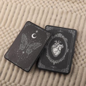 May include: Two dark gray card cases with ornate designs. One features a butterfly with a crescent moon and stars, the other a detailed anatomical heart within a decorative frame. Both have black borders.