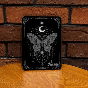 May include: A black rectangular sign with a silver butterfly design, a crescent moon, and stars. The sign has decorative silver borders and the name "Nuvrae" in silver script.