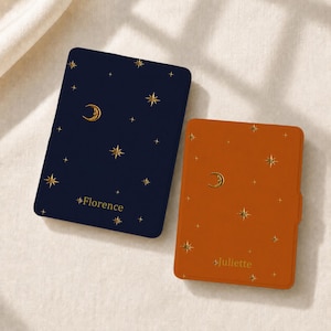 May include: Two rectangular e-reader cases, one dark navy blue and one burnt orange, both featuring a gold foil pattern of stars and a crescent moon. The navy case is personalized with the name Florence, and the orange case with the name Juliette.