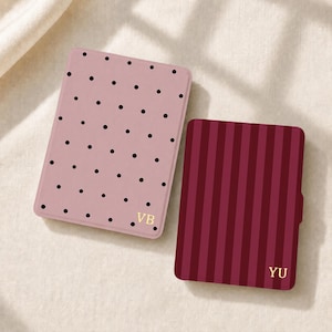 May include: Two rectangular e-reader cases. The left case is dusty pink with a black polka dot pattern and the gold initials VB. The right case features vertical stripes in two shades of burgundy with the gold initials YU in the bottom corner.