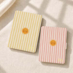 May include: Two rectangular cases with rounded corners, one featuring yellow vertical stripes and the other pink vertical stripes. Each case displays a central illustration of a smiling sun with closed eyes and gold-colored initials FT and DS.