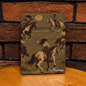 May include: A rectangular plaque featuring an oil-style painting of multiple riders on horseback in a field under a full moon. One rider in the foreground swings a lasso. The scene is rendered in earthy tones of brown, tan, and green with the text Nuvrae.