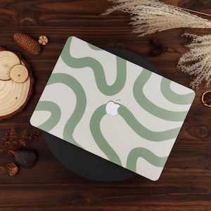 May include: A white laptop case with a green wavy pattern. The case has a cutout for the Apple logo. The design is a modern, abstract pattern. The case is on a black surface.