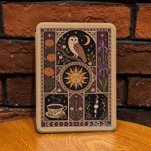 May include: A rectangular e-reader case featuring a detailed illustration of a barn owl on a branch, a sun with a face, a teacup, a key, and moon phases. The design includes orange marigolds and purple irises, all framed in a dark purple border.