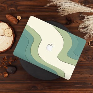 May include: A laptop hard shell case featuring a wavy, layered design in shades of cream, sage green, and dark teal. The case has a central cutout revealing the silver Apple logo. The surface has a smooth finish with curved, abstract lines.