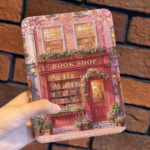 Op de afbeelding: Een rechthoekige hoes met een gedetailleerde illustratie van een charmante boekwinkel. De winkel is geschilderd in tinten rood en roze, met de woorden "BOOK SHOP" boven de ingang. De scène is versierd met bloemen.