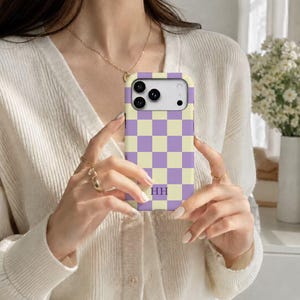 May include: A rectangular phone case featuring a repeating checkerboard pattern in light purple and pale yellow. The case includes a cutout for a multi-lens camera system and displays the letters HH in a purple serif font near the bottom center.