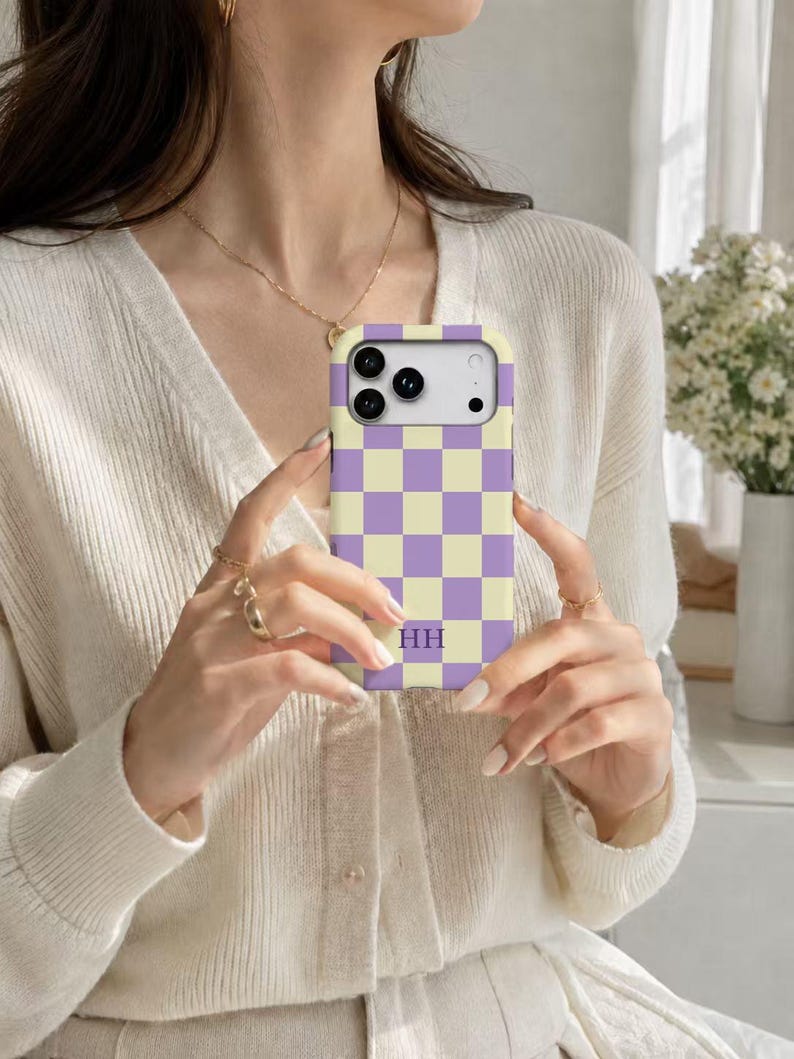 May include: A rectangular phone case featuring a repeating checkerboard pattern in light purple and pale yellow. The case includes a cutout for a multi-lens camera system and displays the letters HH in a purple serif font near the bottom center.