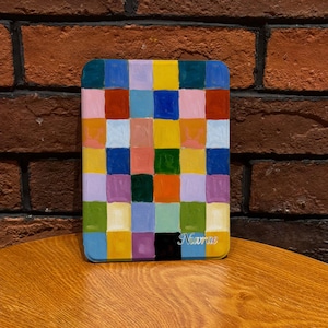 以下が含まれることがあります： 青、黄、赤、緑など様々な色の正方形が格子状に並んだ、長方形のカラフルな絵画。絵画は角が丸く、右下にアーティストのサイン「Narae」が見える。