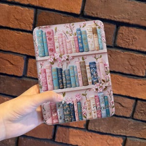 Può includere: Una custodia per telefono con un design a libreria, che mostra file di libri colorati nei toni del rosa, blu e beige. La custodia è decorata con accenti floreali rosa e il testo "Nuvrae".