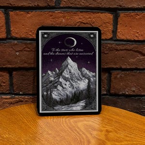 May include: A framed print depicting a mountain range beneath a starry night sky and a crescent moon. The text "To the stars who listen and the dreams that are answered" is written in a decorative script. The print is set within a black frame.