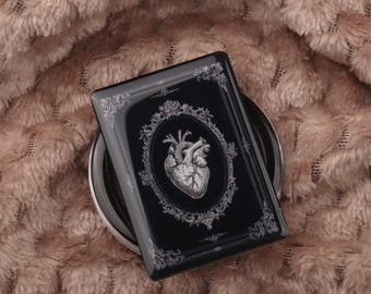 May include: A black compact mirror with a detailed illustration of a human heart in an oval frame. The frame and border have ornate, floral-inspired designs. The mirror rests on a silver base against a soft, textured, peach-colored background.