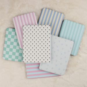 May include: A collection of six rectangular e-reader cases featuring various patterns including pink and white vertical stripes, blue and yellow vertical stripes, a green and white checkerboard, black polka dots on white, and light blue bows.