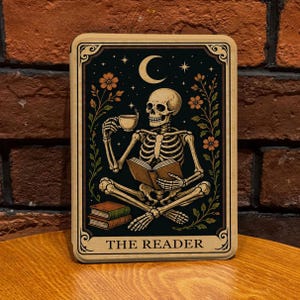 Pode incluir: Uma placa de parede retangular com um esqueleto sentado de pernas cruzadas, lendo um livro aberto enquanto segura uma xícara de chá. A ilustração inclui uma lua crescente, estrelas e vinhas florais num fundo preto, com o texto THE READER.