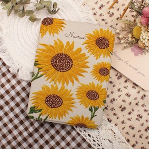 Puede incluir: Una funda rectangular para lector electrónico con fondo beige claro que presenta un patrón de girasoles amarillos con centros marrones. El nombre Nuvrae está impreso en una fuente cursiva negra cerca de la parte superior central de la funda.
