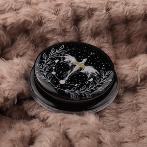 Puede incluir: Un recipiente redondo negro con un diseño celestial. La tapa presenta una ilustración blanca de un dragón y constelaciones sobre un fondo estrellado, enmarcada por una corona floral blanca. El recipiente está sobre una superficie suave y texturizada.