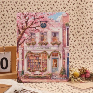 May include: A decorative notebook featuring a bookstore scene with a blooming cherry blossom tree. The illustration includes the word "Bookstore" above the window display of books and the word "Spring" on a sign. The notebook has a light blue door.
