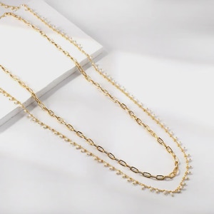 May include: A layered necklace featuring two distinct gold-toned chains. One chain consists of elongated oval links, while the other is a delicate, thinner chain adorned with small, evenly spaced white beads throughout its entire length.