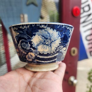May include: A small ceramic bowl featuring a dark blue and cream floral pattern with intricate lattice detailing. The bowl has a wide, flared rim and a circular pedestal base, showcasing a vintage aesthetic with visible signs of age and wear.