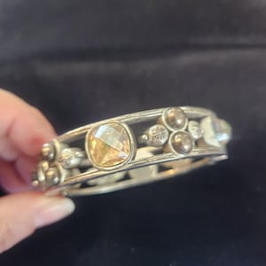 Pu&ograve; includere: Bracciale color argento con una grande gemma ambrata a taglio quadrato al centro. Il bracciale &egrave; decorato con gemme rotonde e trasparenti pi&ugrave; piccole e dettagli metallici decorativi. Lo sfondo &egrave; scuro.