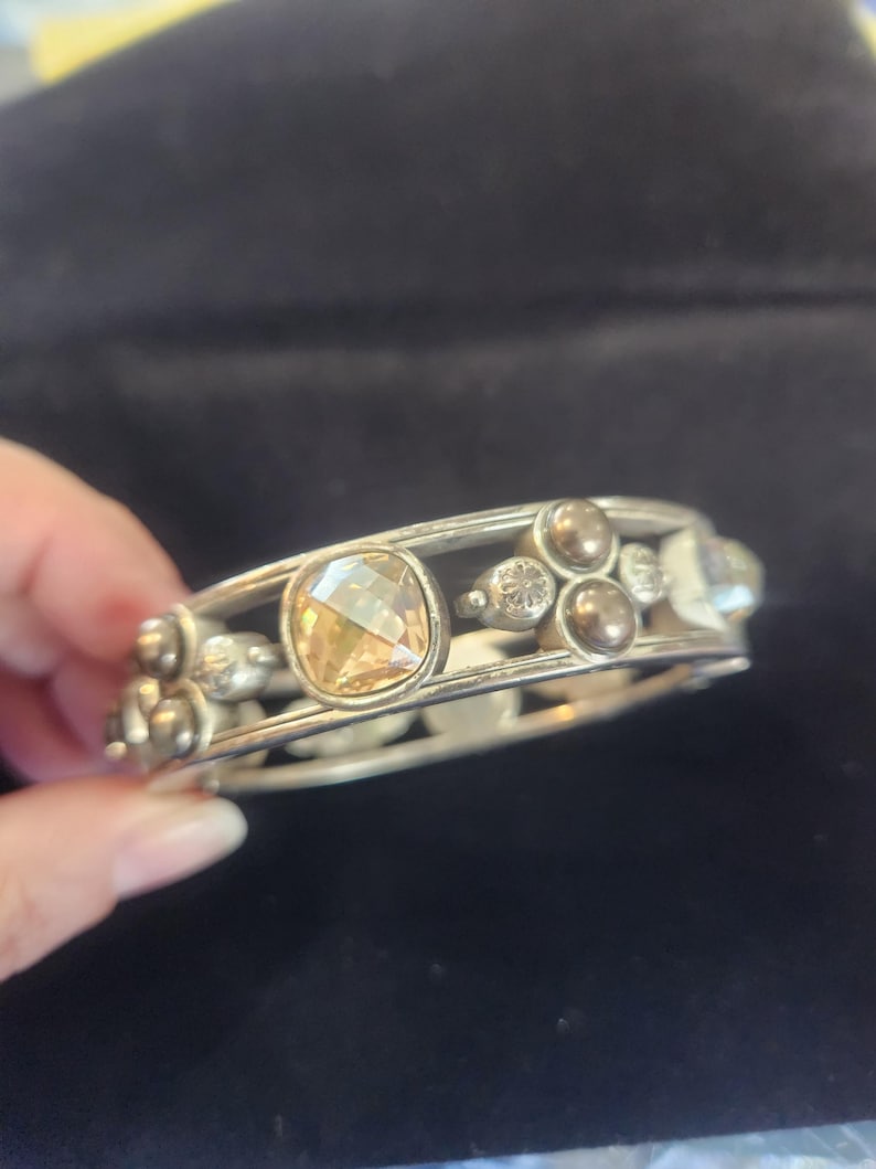 Pu&ograve; includere: Bracciale color argento con una grande gemma ambrata a taglio quadrato al centro. Il bracciale &egrave; decorato con gemme rotonde e trasparenti pi&ugrave; piccole e dettagli metallici decorativi. Lo sfondo &egrave; scuro.