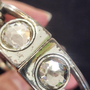 Pu&ograve; includere: Primo piano di un braccialetto argentato con due grandi gemme chiare e sfaccettate. Il braccialetto ha un design rettangolare con le gemme incastonate in cornici circolari. Il metallo mostra segni di usura.