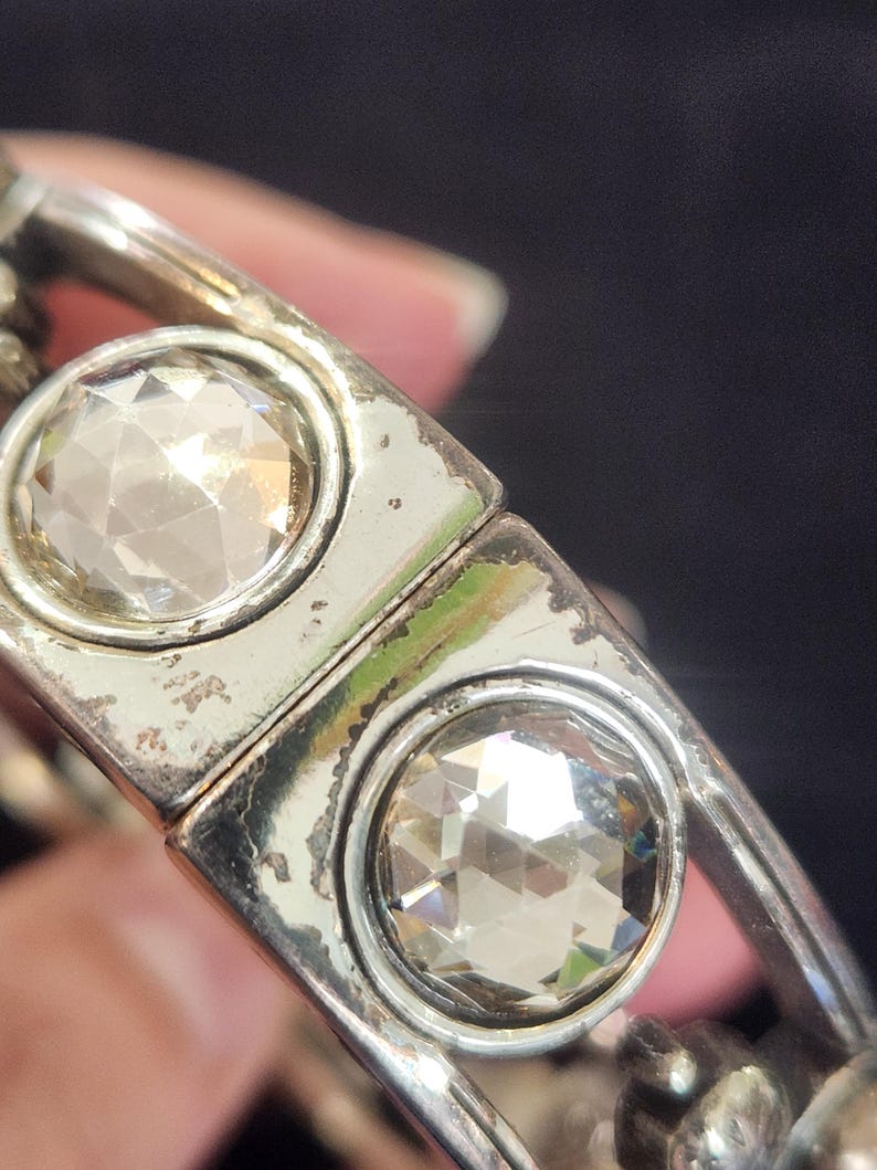 Pu&ograve; includere: Primo piano di un braccialetto argentato con due grandi gemme chiare e sfaccettate. Il braccialetto ha un design rettangolare con le gemme incastonate in cornici circolari. Il metallo mostra segni di usura.