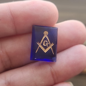 May include: A rectangular, deep blue gemstone featuring a gold-coloured Masonic square and compasses emblem with a central letter G. The stone has a smooth, polished surface and a flat, bevelled edge, showcasing a clear, translucent quality.