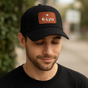 Peut inclure: Casquette de baseball noire avec un patch en cuir marron. Le patch porte le texte "K-Lylo" en blanc, avec une étoile blanche au-dessus. La casquette est en tissu foncé et possède une visière incurvée.