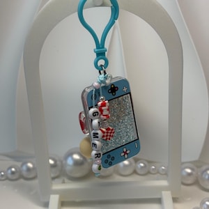 May include: A light blue handheld gaming console keychain featuring a glittery screen, a blue plastic clip, and a dangling charm strand with white letter beads spelling GAMER, accented by red and white checkered and striped cylindrical beads.