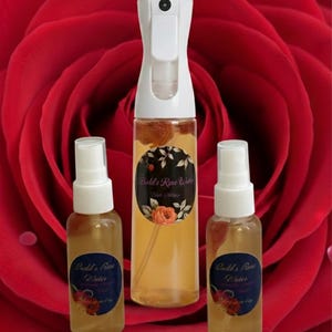 Puede incluir: Tres botellas de spray transparentes llenas de un líquido dorado, etiquetadas como "Budd's Rose Water" y "Cudd's Rose Water". Las botellas están colocadas sobre un fondo de una gran rosa roja.