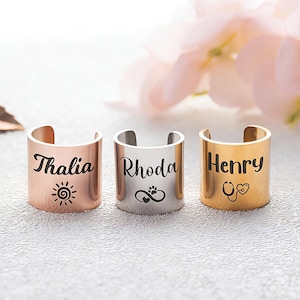 May include: Three open-ended rings in rose gold, silver, and gold. Each ring is engraved with a name and a small symbol: "Thalia" with a sun, "Rhoda" with a paw print, and "Henry" with a stethoscope. The rings are displayed on a textured surface.
