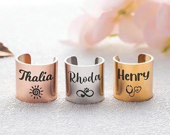 May include: Three open-ended rings in rose gold, silver, and gold. Each ring is engraved with a name and a small symbol: "Thalia" with a sun, "Rhoda" with a paw print, and "Henry" with a stethoscope. The rings are displayed on a textured surface.