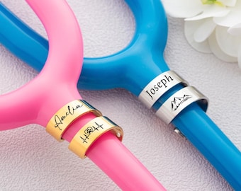 May include: A close-up of two stethoscopes, one pink and one blue. The pink stethoscope has two gold rings engraved with the name "Amelia" and a rose. The blue stethoscope has a silver ring engraved with the name "Joseph" and a mountain scene.