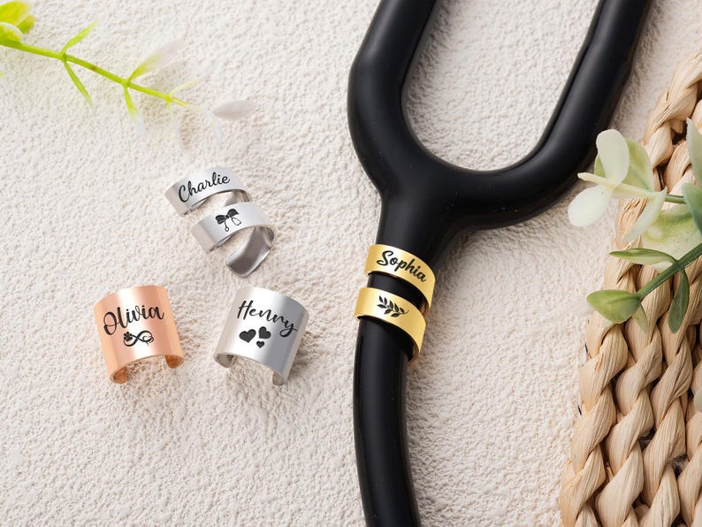 May include: A collection of personalised metal stethoscope identification tags in silver, gold, and rose gold tones. The tags feature engraved names including Charlie, Olivia, Henry, and Sophia, alongside small icons like hearts, a bow, and a leaf.