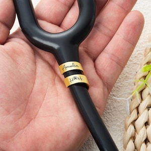 May include: A black stethoscope tubing section featuring two gold-coloured metal rings. The top ring is engraved with the name Amelia in a script font, and the bottom ring displays a heartbeat line graphic with a small paw print symbol in the centre.