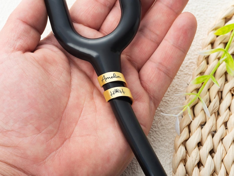 May include: A black stethoscope tubing section featuring two gold-coloured metal rings. The top ring is engraved with the name Amelia in a script font, and the bottom ring displays a heartbeat line graphic with a small paw print symbol in the centre.