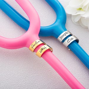 May include: A bright pink and a bright blue tuning fork, each featuring two metallic rings around the handle. The pink fork has gold-toned rings engraved with the name Amelia and a heart-beat symbol. The blue fork has silver-toned rings engraved with the name Joseph and a mountain icon.