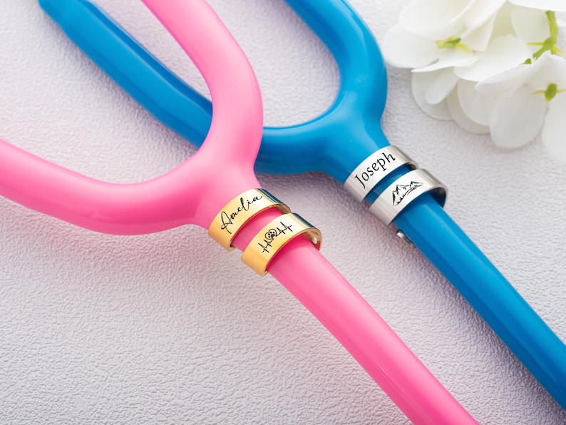 May include: A bright pink and a bright blue tuning fork, each featuring two metallic rings around the handle. The pink fork has gold-toned rings engraved with the name Amelia and a heart-beat symbol. The blue fork has silver-toned rings engraved with the name Joseph and a mountain icon.