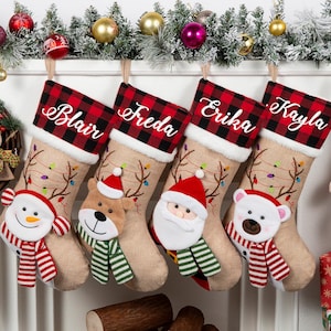 May include: Four tan hessian Christmas stockings featuring a red and black buffalo check cuff with white embroidered names Blair, Freda, Erika, and Kayla. Each stocking displays a plush character: a snowman, a reindeer, Santa, and a polar bear.