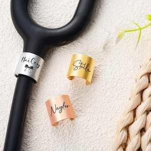 May include: A black crutch handle featuring a silver identification band engraved with the name Hailey and a bow icon, alongside two separate metal bands in gold and rose gold engraved with the names Stella and Naylor respectively.