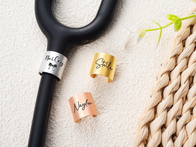 May include: A black crutch handle featuring a silver identification band engraved with the name Hailey and a bow icon, alongside two separate metal bands in gold and rose gold engraved with the names Stella and Naylor respectively.