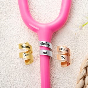 May include: A bright pink stethoscope tubing section adorned with three spiral metal identification tags. The gold tag is engraved with Sarah and a stethoscope icon, the silver tag with Bailey and hearts, and the rose gold tag with Kristin and a paw print.